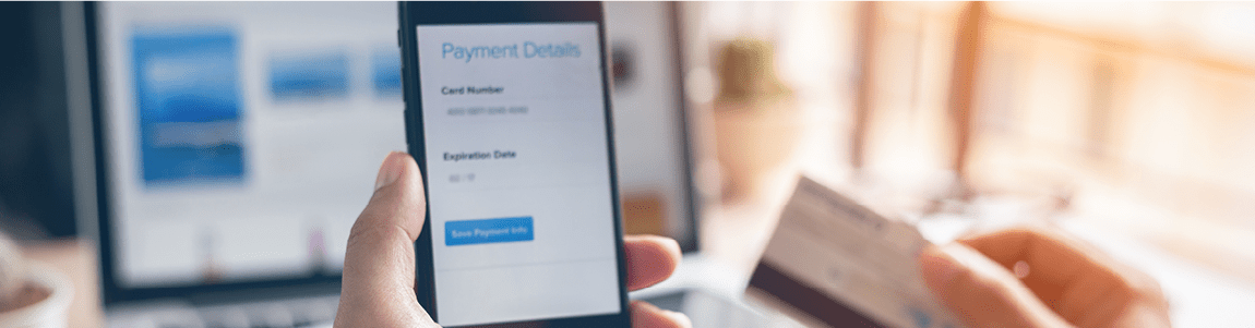 A person holds a smartphone displaying a payment details form while typing from a credit card, with a blurred laptop screen in the background.