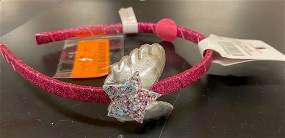 A pink glittery headband with a metallic silver seashell and a multicolored glitter star adornment, placed on a black surface with attached price tags.