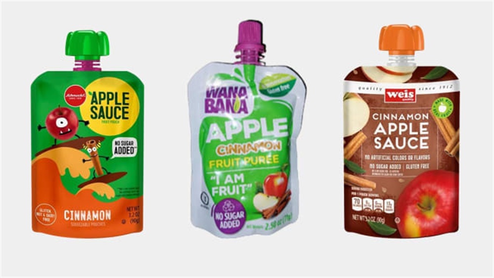 Three apple cinnamon puree pouches with vibrant packaging. Left: green cap, cartoon apple; center: purple cap, fruit imagery; right: orange cap, cinnamon sticks.