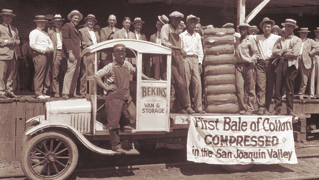 Historical photo of men on a truck with bale of cotton and sign reading first bale of cotton compressed in the San Joaquin Valley