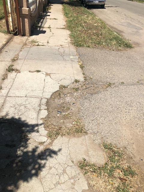 View of a narrow, damaged sidewalk with extensive cracking, broken concrete, and an uneven surface where the walkway meets the edge of the pavement.