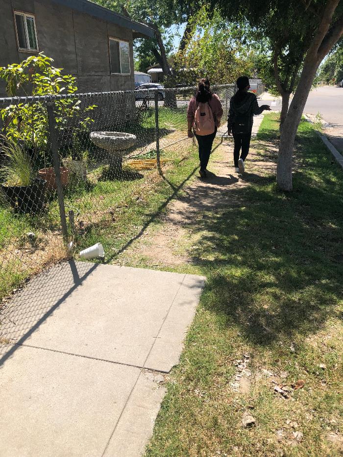 View of a sidewalk segment where the paved walkway ends and students are walking on a narrow dirt path beside a chain-link fence. The concrete sidewalk is only partially constructed, and grass and uneven ground fill the gap between sidewalk sections.