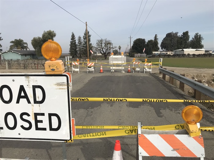 Photo of the Houghton Canal Bridge on N. Chateau Fresno Ave. The bridge is closed with multiple barricades, orange warning lights, cones, and caution tape blocking the roadway. Guardrails line both sides of the bridge. Beyond the closure, the road continues past fields, trees, and nearby buildings.