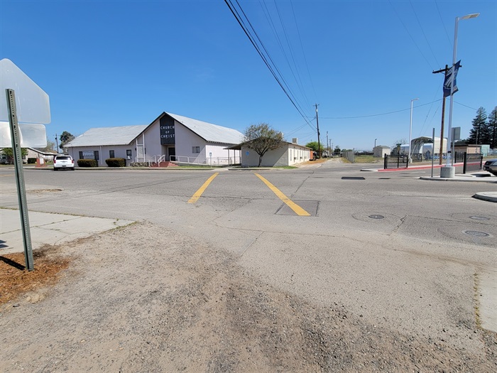 Facing northeast across Quince near the northwest entrance of the elementary school. There is rough pavement leading to an intersection with two yellow painted crosswalk markings. Across the street is a building with a sign above the entrance that reads ‘Church of Christ.’